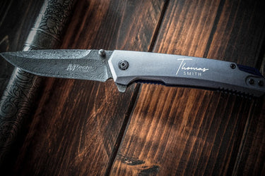 Damascus-pattern folding knife, open, with a name in script on the anodized handle