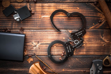 Black novelty handcuffs laid in a heart shape, a short phrase engraved on each cuff